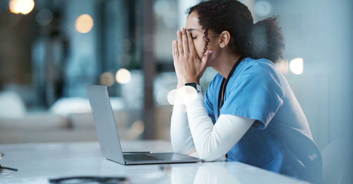 nurse sits at laptop with hands over her face and appears stressed